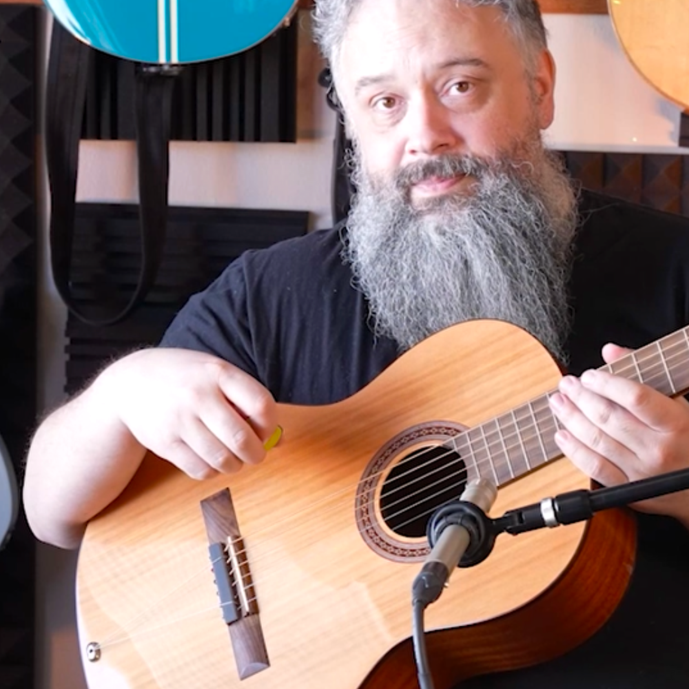 Man playing an acoustic guitar with a TurboStrand in a music studio with guitars and equipment in the background.